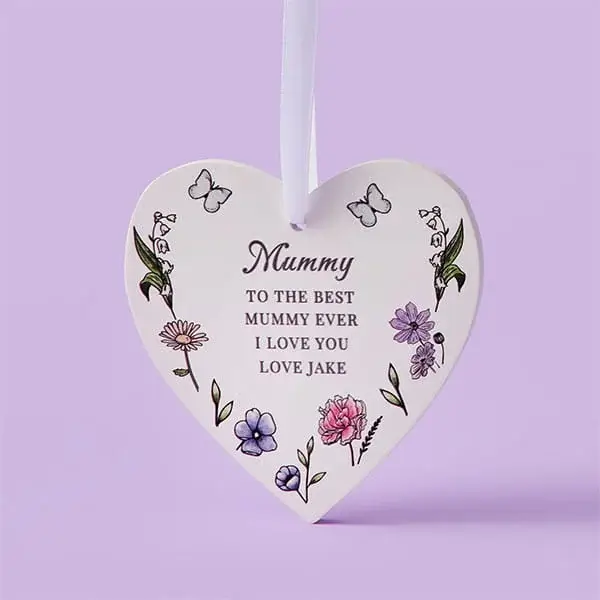 A heart-shaped ceramic hanging decoration with floral illustrations and a heartfelt message for a mum, shown against a pastel purple background.